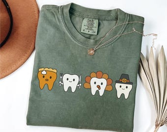 May include: An olive green t-shirt featuring a graphic of four cartoon teeth dressed as Thanksgiving foods. The teeth are designed as a slice of pie, a tooth with a fork and knife, a turkey, and a pilgrim. A gold necklace is draped across the shirt.