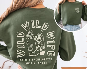 May include: Military green sweatshirt with a white graphic that says "WILD WIFE" with a disco ball, cowboy boot, and ring. The text "KATIE'S BACHELORETTE AUSTIN, TEXAS" is also printed on the sweatshirt.