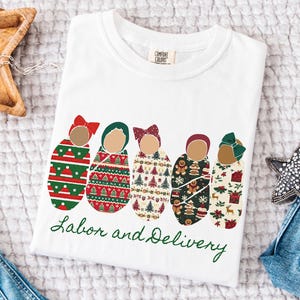 Peut inclure: T-shirt blanc avec un motif festif repr&eacute;sentant cinq b&eacute;b&eacute;s emmaillot&eacute;s, chacun orn&eacute; d'un n&oelig;ud et envelopp&eacute; dans des motifs sur le th&egrave;me des f&ecirc;tes. Les mots "Labor and Delivery" sont &eacute;crits en vert.