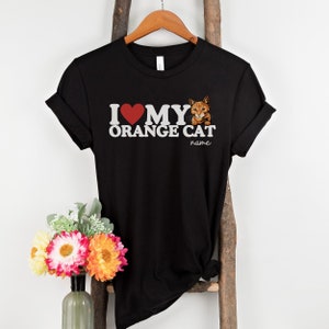 Op de afbeelding: Zwart T-shirt met een witte afbeelding waarop staat "Ik hou van mijn oranje kat" met een rood hart en een afbeelding van een oranje kat. De tekst "name" staat onder de afbeelding.