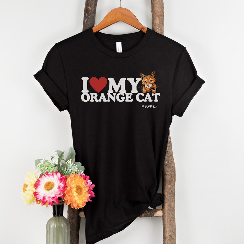 Op de afbeelding: Zwart T-shirt met een witte afbeelding waarop staat "Ik hou van mijn oranje kat" met een rood hart en een afbeelding van een oranje kat. De tekst "name" staat onder de afbeelding.
