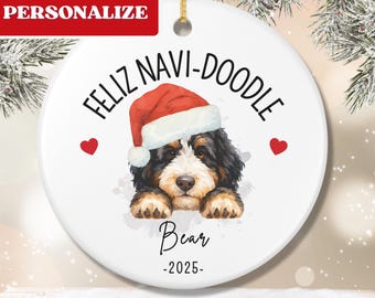 Op de afbeelding: Ronde witte keramische kerstversiering met een illustratie van een Bernedoodle met een kerstmuts. De versiering heeft de tekst "FELIZ NAVI-DOODLE" en "Bear -2025". Twee rode harten flankeren de hond.