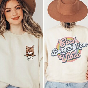 Può includere: Felpa color crema con grafica di gatto Bengal e il nome "Spice" sotto. Il retro della felpa mostra il testo "Cool Bengal Mom Vibes" in un design retrò con un arcobaleno.