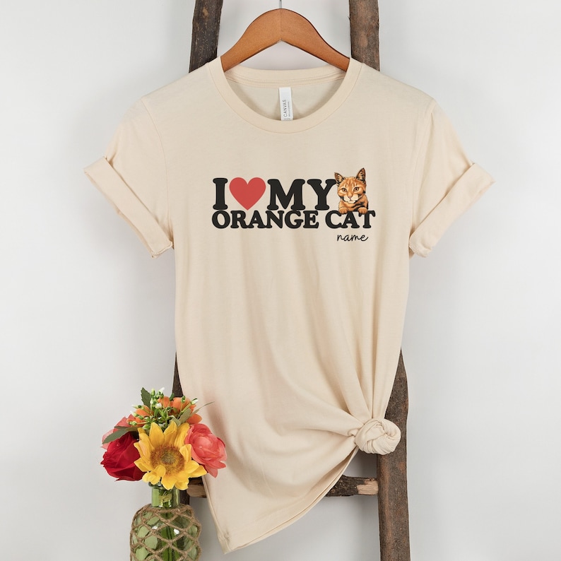 Op de afbeelding: Een beige T-shirt met de tekst "Ik hou van mijn oranje kat" en een rood hart. Een afbeelding van een oranje kat bevindt zich in de rechterbovenhoek van het shirt. De tekst "name" staat onder de kat.