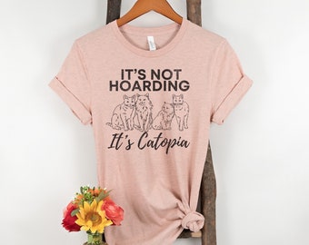 Puede incluir: Una camiseta rosa con un gráfico de cinco gatos y el texto "It's not hoarding It's Catopia".