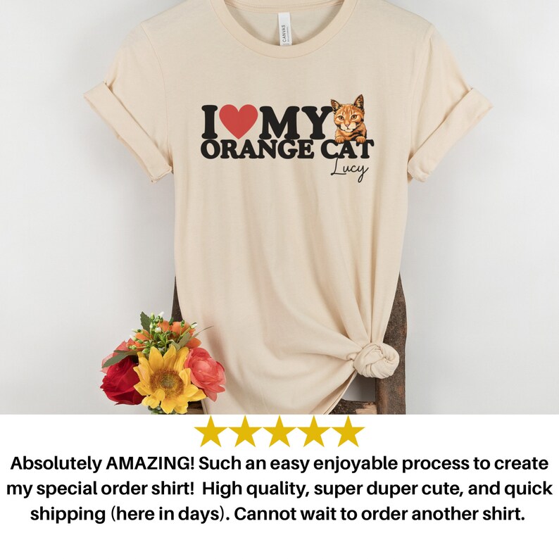 Op de afbeelding: Een beige T-shirt met een zwarte afbeelding waarop staat "Ik hou van mijn oranje kat" met een rood hart en een afbeelding van een oranje kat. De naam van de kat, "Lucy", staat onder de afbeelding.