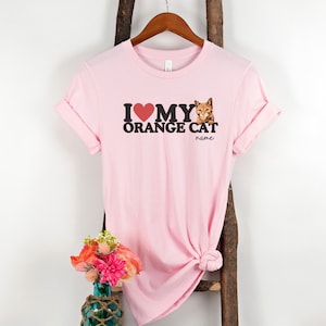 Op de afbeelding: Roze T-shirt met de tekst "Ik hou van mijn oranje kat" en een afbeelding van een oranje kat. De tekst "name" staat onder de kat.