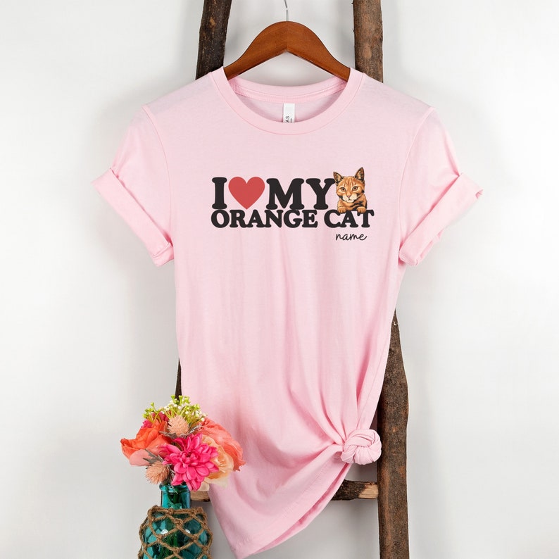 Op de afbeelding: Roze T-shirt met de tekst "Ik hou van mijn oranje kat" en een afbeelding van een oranje kat. De tekst "name" staat onder de kat.