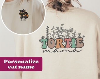 May include: A beige sweatshirt with a personalized cat name and a graphic of a cat with the text "Tortie mama" and floral accents.