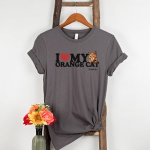 Op de afbeelding: Grijs T-shirt met een rood hart, zwarte tekst die "Ik hou van mijn oranje kat" zegt en een afbeelding van een oranje kat. De tekst "naam" staat onder de kat.