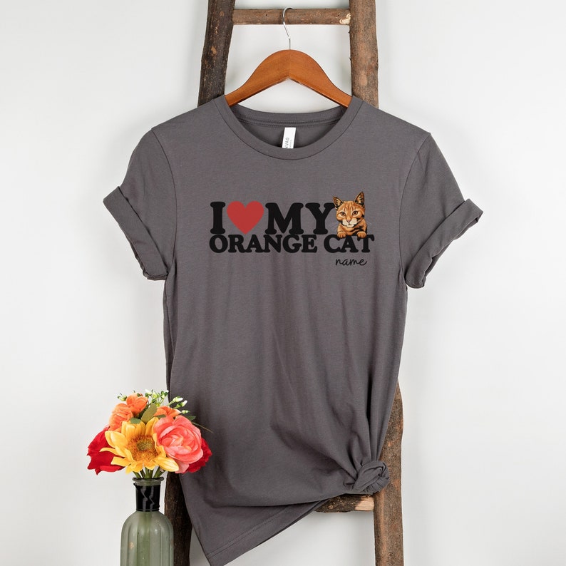Op de afbeelding: Grijs T-shirt met een rood hart, zwarte tekst die "Ik hou van mijn oranje kat" zegt en een afbeelding van een oranje kat. De tekst "naam" staat onder de kat.
