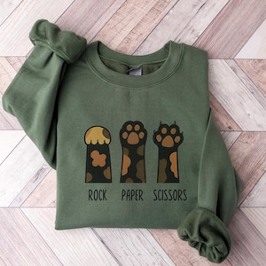 Può includere: Una felpa verde oliva con tre impronte stilizzate di zampe di gatto in marrone e beige, che rappresentano sasso, carta e forbici. Le parole "ROCK PAPER SCISSORS" sono stampate in nero sotto. La felpa ha le maniche lunghe.