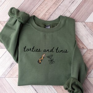 Può includere: Una felpa verde oliva con la scritta ricamata "totties and tinis". Il design include un'immagine a cartoni animati di una zampa di gatto e un bicchiere da martini. La felpa ha un girocollo e maniche lunghe.