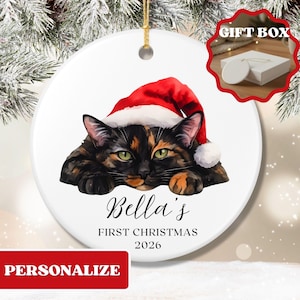 May include: A round white ceramic ornament featuring a tortoiseshell cat wearing a red Santa hat. The text reads Bella's FIRST CHRISTMAS 2026. The ornament has a gold hanging string and is displayed against a snowy pine branch background.
