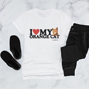 Op de afbeelding: Wit T-shirt met een rood hart en de tekst "Ik hou van mijn oranje kat" met een afbeelding van een oranje kat. De tekst "name" staat onder de tekst.
