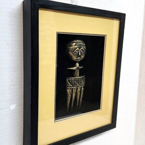 May include: A framed artwork featuring a gold-coloured sculpture of a stylised human figure with a crown and a comb-like object. The sculpture is set against a black background.