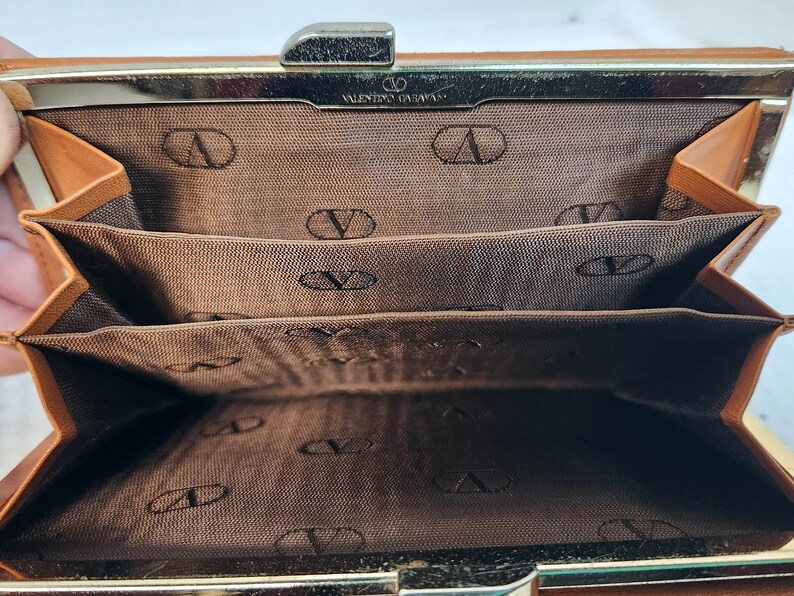 May include: A brown leather wallet with a gold clasp and multiple card slots. The interior is lined with brown fabric and features the Valentino Garavani logo.