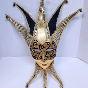 May include: A gold and black jester mask with a pointed hat and bells. The mask has a detailed design with swirls and a black and gold pattern.