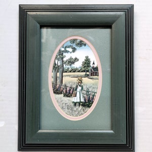 以下が含まれることがあります： 田舎の風景の中に、白いドレスと麦わら帽子をかぶった少女が赤い校舎に向かって歩いている絵が描かれた額装されたプリント。絵はピンクと緑のマットが付いた楕円形のフレームに収められている。