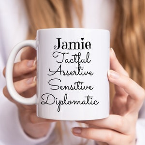 May include: White ceramic mug with a black design that says "Jamie Tacftul Assertive Sensitive Diplomatic" with a small heart after the name.