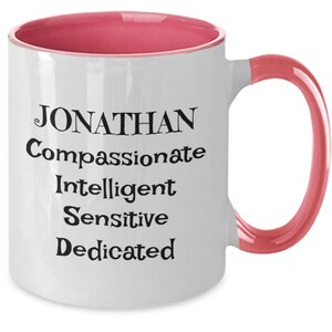 May include: White ceramic coffee mug with a pink rim and handle. The mug has black text that reads "JONATHAN Compassionate Intelligent Sensitive Dedicated".