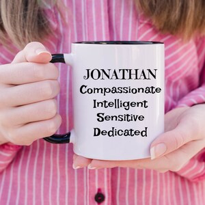 May include: White ceramic mug with a black handle and black rim. The mug has the name "JONATHAN" printed in black, followed by the words "Compassionate", "Intelligent", "Sensitive", and "Dedicated".
