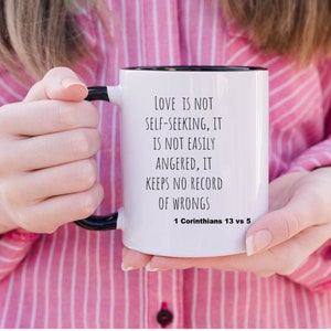 Puede incluir: Taza de cerámica blanca con borde y asa negros. La taza tiene texto negro que dice "El amor no es egoísta, no se irrita fácilmente, no guarda rencor. 1 Corintios 13 vs 5".