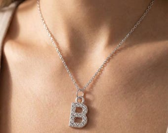 Può includere: Una collana color argento con un ciondolo a forma di lettera "B". Il ciondolo è incastonato con piccoli cristalli trasparenti ed è sospeso a una catena sottile. La collana è mostrata su uno sfondo neutro.