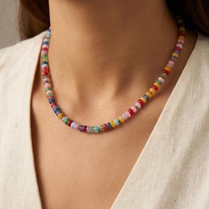 以下が含まれることがあります： 赤、ピンク、黄色、青、緑など、さまざまな色の小さな丸いビーズで作られたカラフルなネックレス。 ネックレスは首にかけられ、クリーム色のトップスの上にあります。