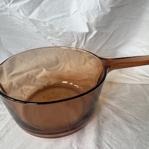 Pode incluir: Uma panela de vidro transparente na cor âmbar, com um cabo longo e plano de um lado e uma pequena alça moldada no lado oposto. O recipiente possui uma abertura larga e uma base lisa e curva, fabricado em vidro resistente ao calor.