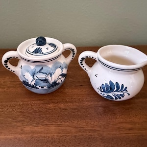 May include: A white ceramic sugar bowl with a lid and a matching creamer pitcher, both decorated with blue hand-painted designs. The sugar bowl features a windmill scene, while the creamer displays a leaf pattern. Both pieces have blue-dotted handles.