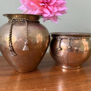 Può includere: Due vasi in ottone lucido dalla forma arrotondata e con un bordo largo. Ogni contenitore presenta un decoro in corda di ottone intrecciata avvolto attorno al collo con dettagli a nappa pendenti. Un fiore di dalia rosa brillante riposa nel vaso più alto.