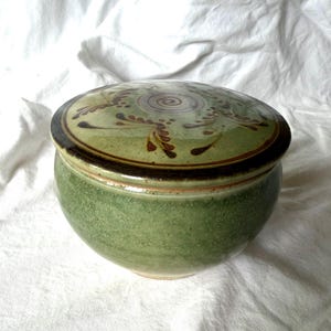 May include: A round ceramic lidded jar with a glossy green glaze and a speckled texture. The lid features a central spiral design surrounded by brown leaf-like patterns. The rim of the lid is dark brown, and the base of the jar is rounded.