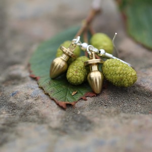 Puede incluir: Un par de pendientes dorados con un diseño alargado único. Los pendientes se muestran sobre una hoja verde con pequeñas piñas verdes texturizadas. El fondo es una superficie de piedra texturizada y borrosa.