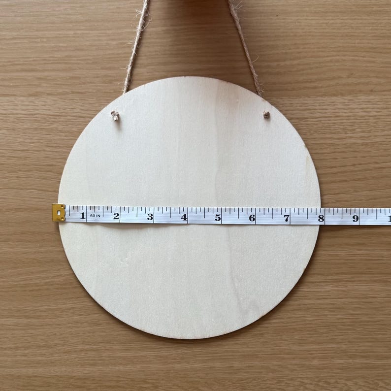 Pode incluir: Uma placa redonda de madeira inacabada com gr&atilde;o natural, com aproximadamente 20 cm de di&acirc;metro. Uma fita m&eacute;trica &eacute; colocada sobre a placa e uma corda de juta &eacute; presa para pendurar.