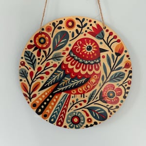 Pode incluir: Decora&ccedil;&atilde;o de parede redonda em madeira com um design estilizado de p&aacute;ssaro. O p&aacute;ssaro &eacute; predominantemente vermelho, azul e laranja, rodeado por motivos florais e foliares. Uma al&ccedil;a de barbante natural &eacute; anexada para pendurar.