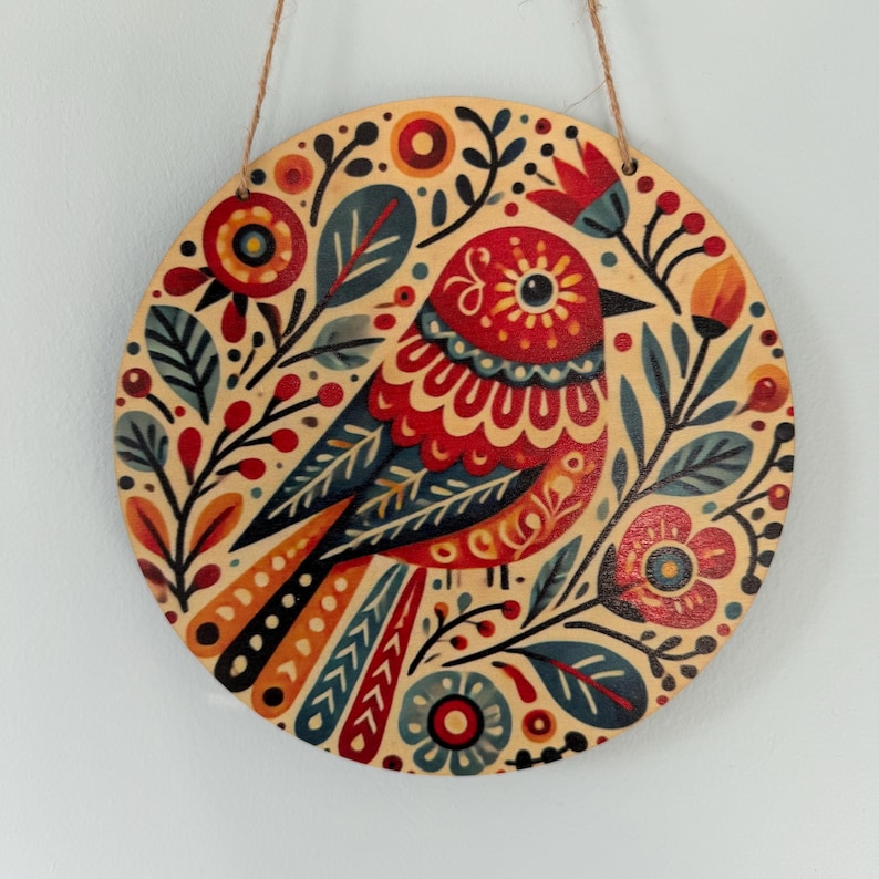 Pode incluir: Decora&ccedil;&atilde;o de parede redonda em madeira com um design estilizado de p&aacute;ssaro. O p&aacute;ssaro &eacute; predominantemente vermelho, azul e laranja, rodeado por motivos florais e foliares. Uma al&ccedil;a de barbante natural &eacute; anexada para pendurar.