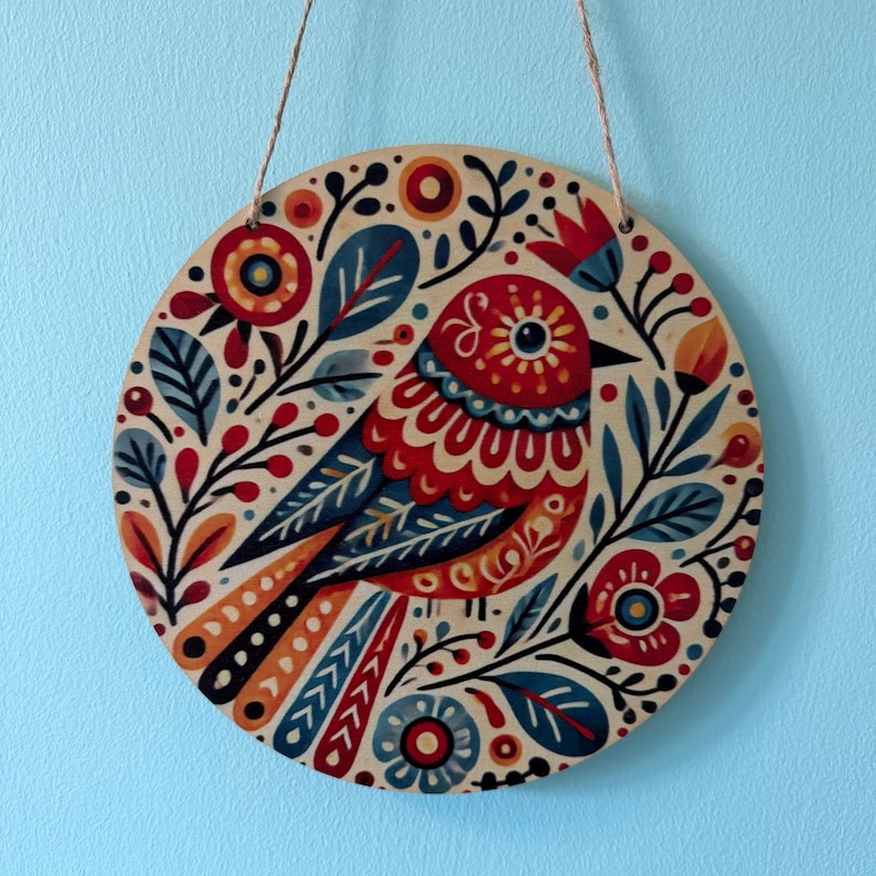 Pode incluir: Arte de parede redonda em madeira com um design de p&aacute;ssaro colorido. O p&aacute;ssaro &eacute; principalmente vermelho, azul e laranja, rodeado por motivos florais e de folhas. Uma corda de juta &eacute; fixada para pendurar. Fundo azul claro.