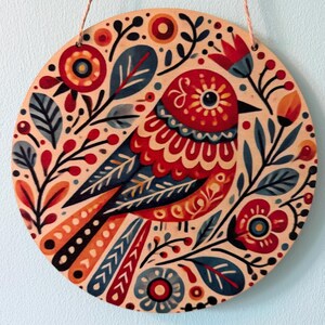 Pode incluir: Arte de parede redonda em madeira com um design de p&aacute;ssaro colorido. O p&aacute;ssaro &eacute; principalmente vermelho, azul e laranja, rodeado por motivos florais e de folhas. A obra de arte est&aacute; sobre um fundo bege claro e tem uma corda para pendurar.