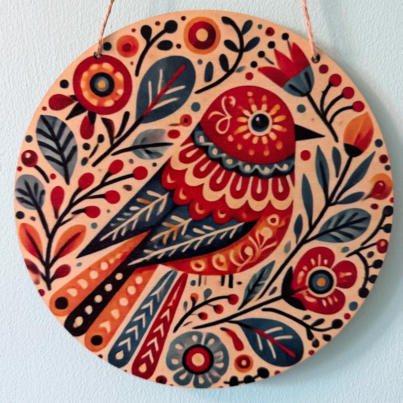 Pode incluir: Arte de parede redonda em madeira com um design de p&aacute;ssaro colorido. O p&aacute;ssaro &eacute; principalmente vermelho, azul e laranja, rodeado por motivos florais e de folhas. A obra de arte est&aacute; sobre um fundo bege claro e tem uma corda para pendurar.