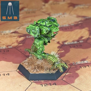 May include: A green miniature mech robot with red eyes stands on a hexagonal base. The robot has detailed armour and weaponry. The base is painted to resemble a cracked desert landscape. The SMB logo is in the upper left corner.