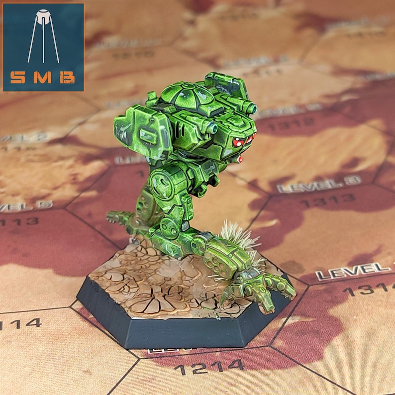 May include: A green miniature mech robot with red eyes stands on a hexagonal base. The robot has detailed armour and weaponry. The base is painted to resemble a cracked desert landscape. The SMB logo is in the upper left corner.