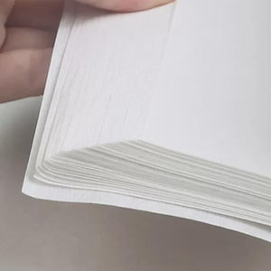 Könnte beinhalten: Ein Stapel leerer, weißer, rechteckiger Papierbögen mit glatter, matter Textur. Die Kanten des Papiers sind sauber ausgerichtet und zeigen die Dicke der mehreren Schichten aus dünnen, undurchsichtigen weißen Blättern, die in einem Stapel liegen.