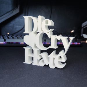 Puede incluir: Una escultura blanca impresa en 3D con las palabras "Die Cry Hate" apiladas verticalmente. Un corazón está entre "Die" y "Cry". La escultura está sobre una superficie oscura.