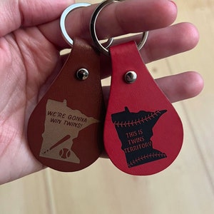 May include: Two leather keychains shaped like the state of Minnesota. One is brown with the text "We're gonna win Twins!" and a baseball bat and ball. The other is red with the text "This is Twins Territory" and a baseball stitch design.