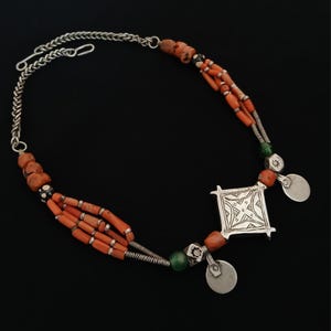 May include: A handcrafted necklace featuring orange coral beads, silver chain, and a square pendant with an etched design. The necklace includes small silver disc accents and green beads, creating a unique, ethnic jewelry piece.