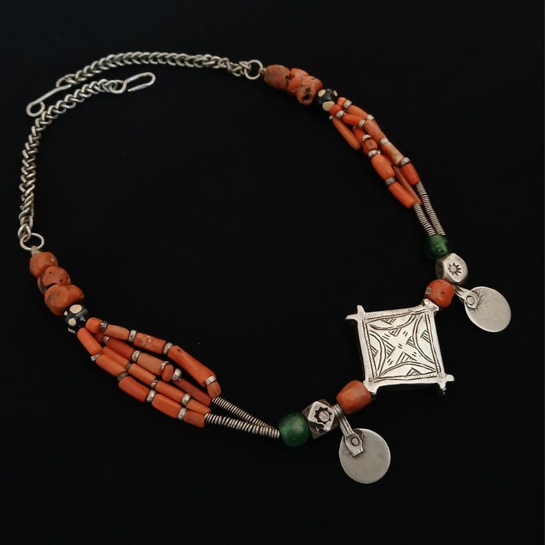 May include: A handcrafted necklace featuring orange coral beads, silver chain, and a square pendant with an etched design. The necklace includes small silver disc accents and green beads, creating a unique, ethnic jewelry piece.