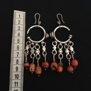 May include: A pair of silver-toned dangle earrings with a crescent-shaped top and dangling coral-colored beads. The earrings have a hook closure and are displayed against a black background.