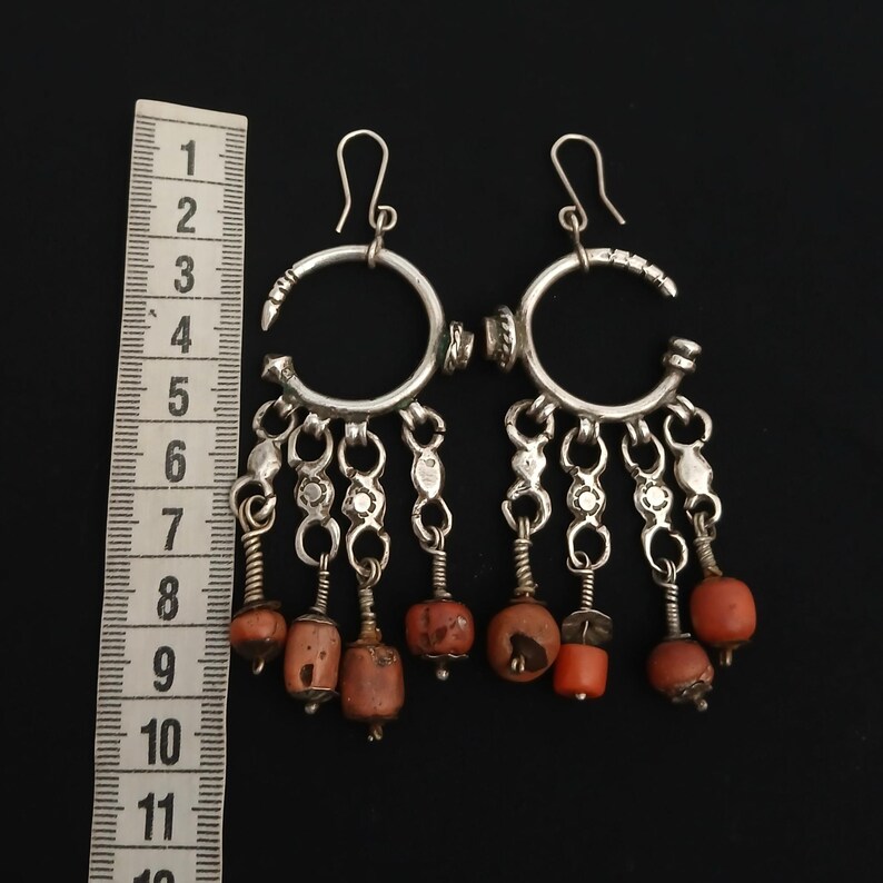 May include: A pair of silver-toned dangle earrings with a crescent-shaped top and dangling coral-colored beads. The earrings have a hook closure and are displayed against a black background.