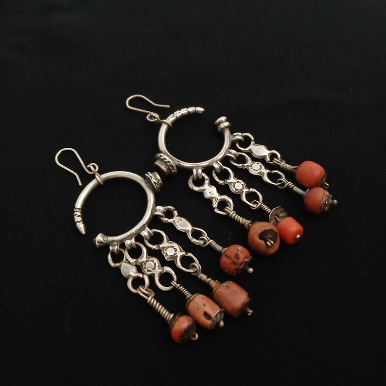 May include: A pair of silver-toned earrings with a circular design and dangling chains. Each earring features small, orange-red beads and decorative metalwork. The earrings are displayed against a black background.